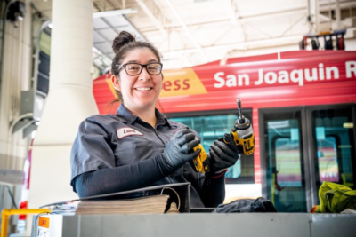 Female RTD mechanic holding onto a power drill. She is standing in front of an RTD bus.