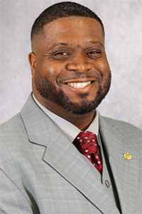 Portrait of Aaron Edwards smiling, wearing a light gray suit jacket, gray vest, white dress shirt, and a red patterned tie, photographed against a neutral gray studio background.