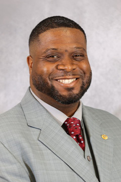Board Member Aaron Edwards wearing a light gray suit, white shirt, and red patterned tie, smiling against a gray studio background.