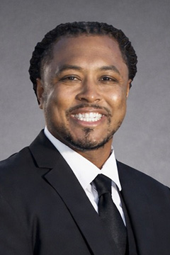 Board Member Derek Graves wearing a black suit, white shirt, and black tie, smiling against a dark gray studio background.