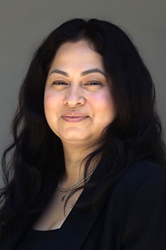 Board Member Geneva Moorad wearing a black blazer and necklace, smiling against a neutral gray background.